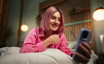 Ragazza sul letto con smartphone in mano che sorride mentre invia un messaggio della buonanotte