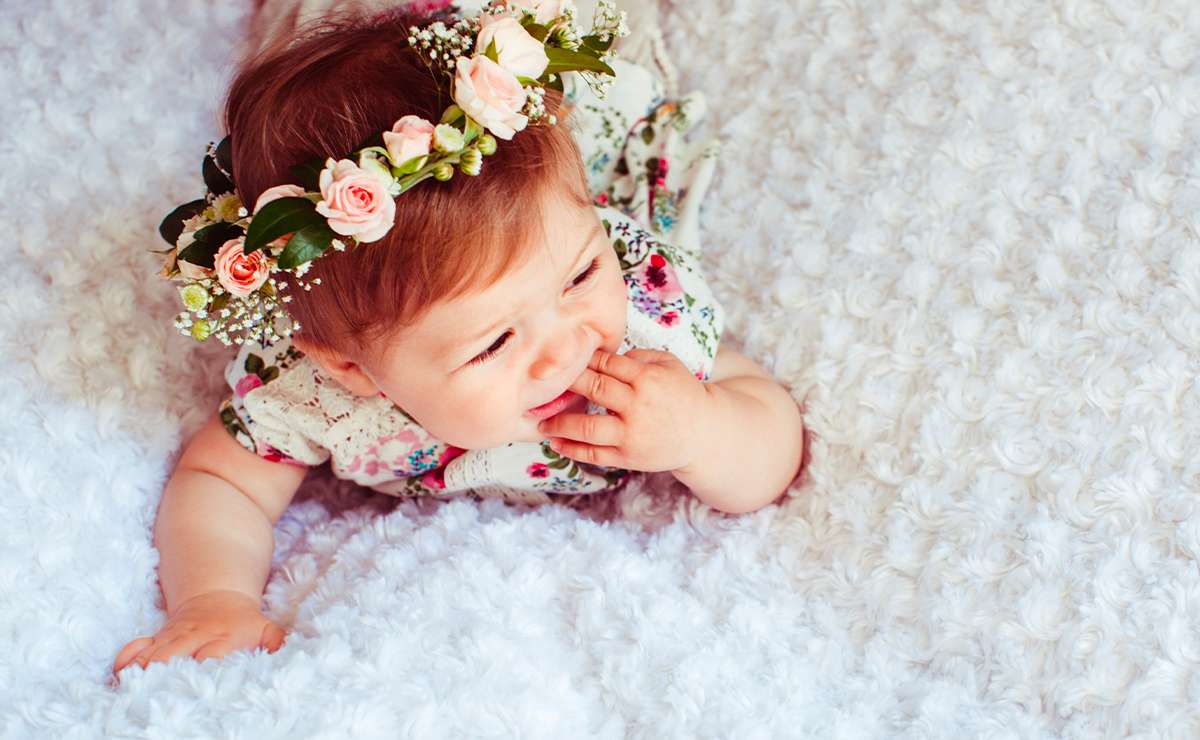 Bambina sdraiata su coperta morbida bianca con corona di fiori rosa e vestitino floreale
