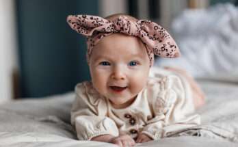 Bambina che sorride sdraiata sul letto con fascia rosa a fiocco e vestitino chiaro