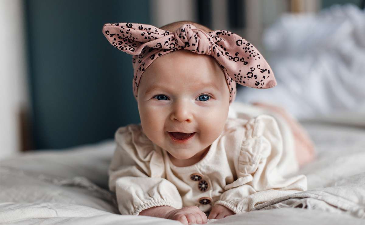 Bambina che sorride sdraiata sul letto con fascia rosa a fiocco e vestitino chiaro
