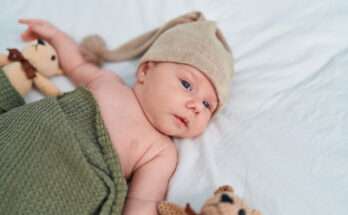 Neonato disteso sul letto con cappellino beige e copertina verde, affiancato da pupazzi, immagine dedicata ai nomi maschili italiani con la Z