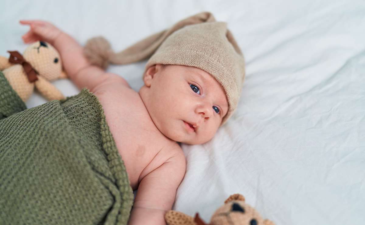 Neonato disteso sul letto con cappellino beige e copertina verde, affiancato da pupazzi, immagine dedicata ai nomi maschili italiani con la Z