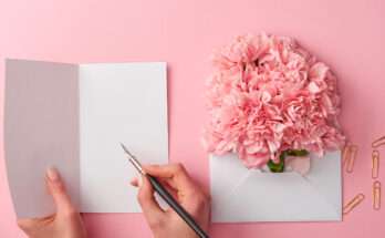 Mano che scrive un messaggio di auguri su un biglietto bianco, accanto a una busta aperta con fiori rosa