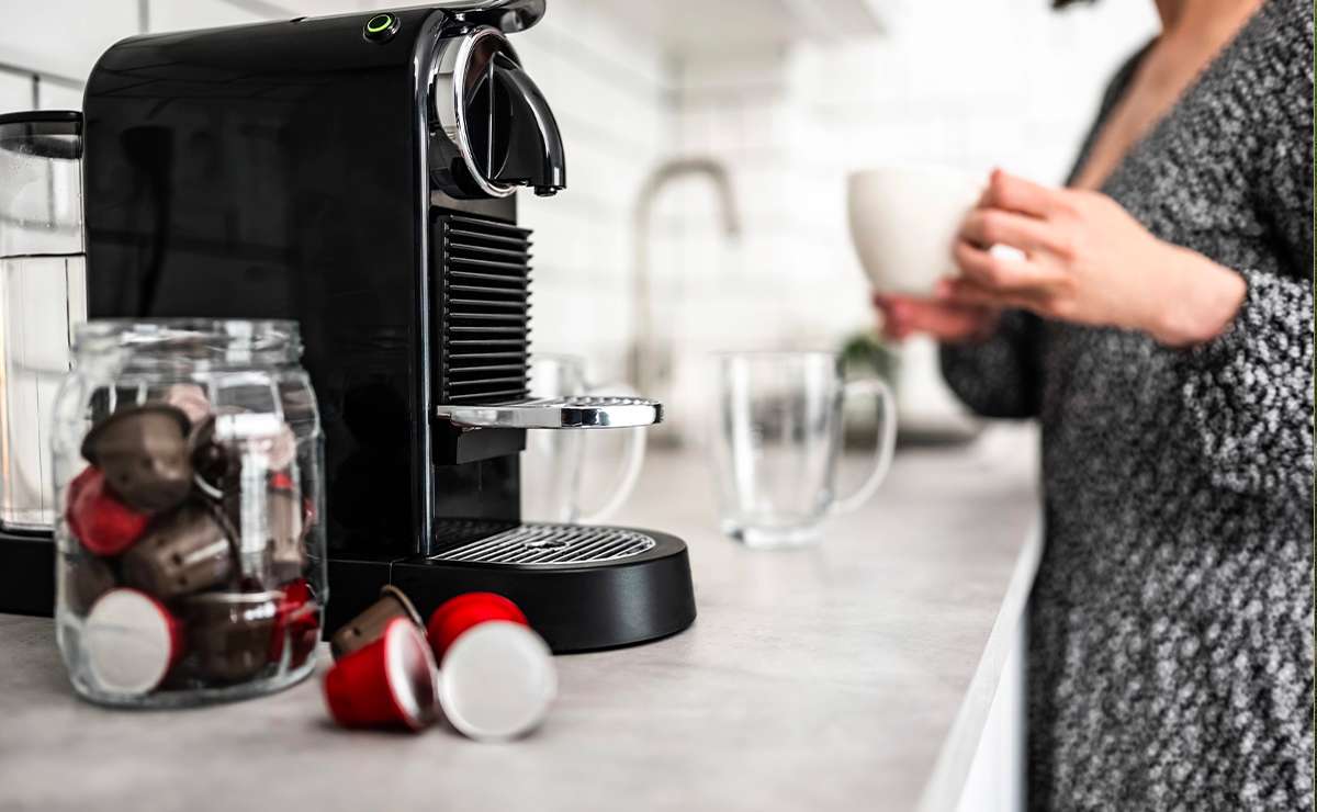 Macchina da caffè a capsule su piano cucina con barattolo di cialde e donna che sorseggia una tazza