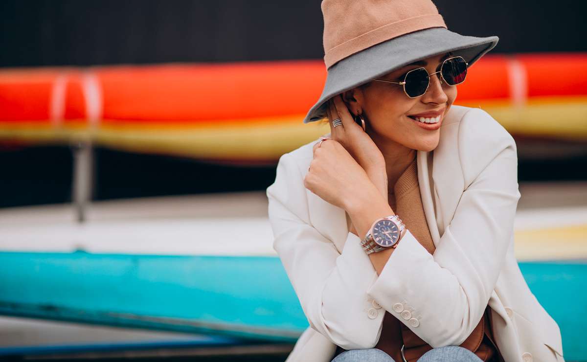 Donna con blazer chiaro, cappello beige e occhiali da sole indossa un orologio da polso elegante