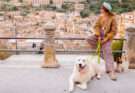 Donna con cappello e cane bianco seduti su un belvedere con vista su un borgo italiano