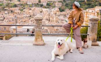 Donna con cappello e cane bianco seduti su un belvedere con vista su un borgo italiano