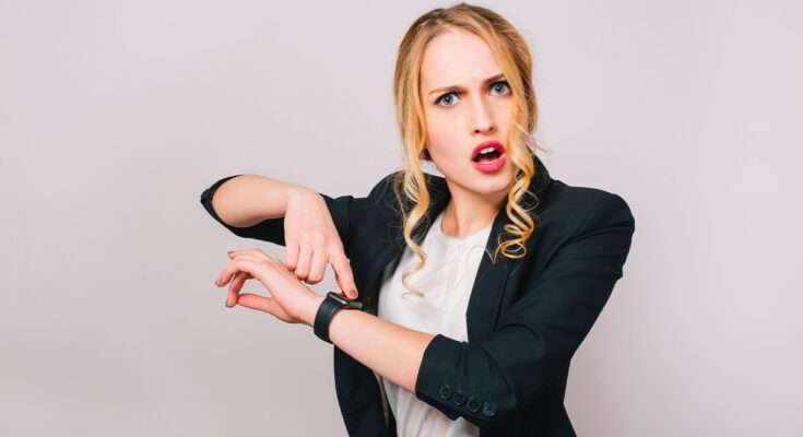 donna bionda indica l'ora sull'orologio da polso con espressione stupita
